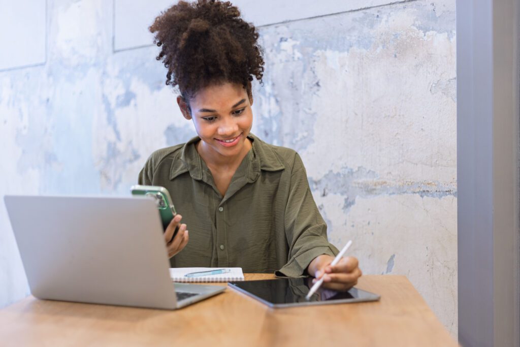 Happy young African American woman using digital smartphone and laptop sitting at wood desk